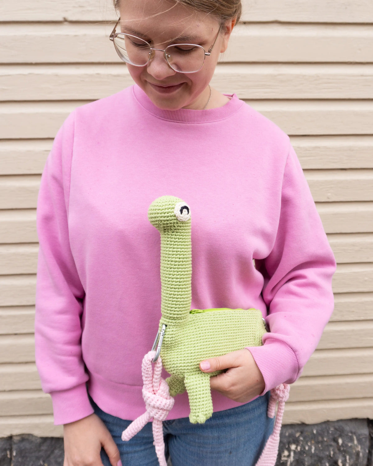 Person wearing a green crocheted brachiosaurus dinosaur bag and a pink hoodie.