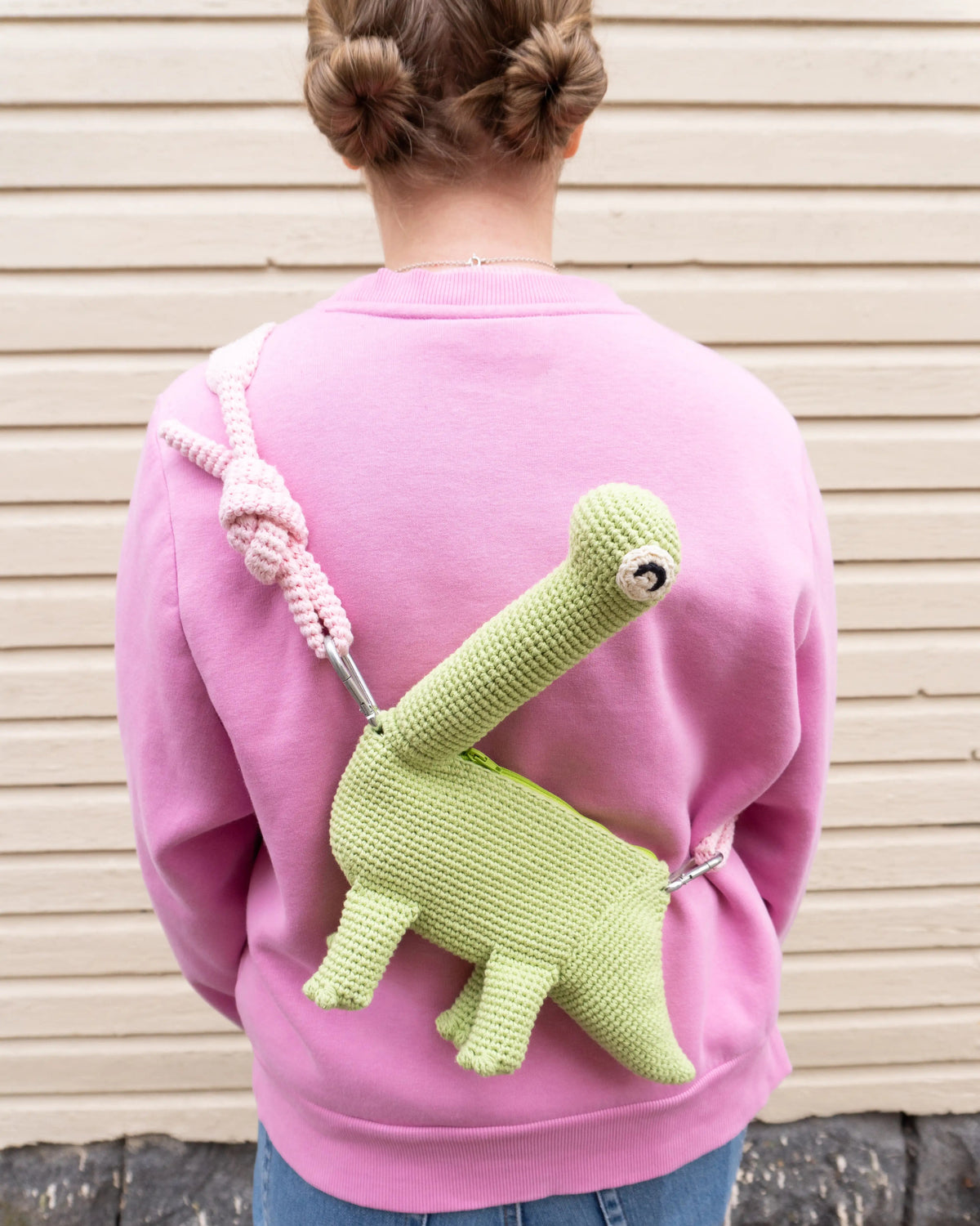 Person wearing a green crocheted brachiosaurus dinosaur bag and a pink hoodie.