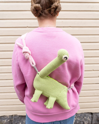 Person wearing a green crocheted brachiosaurus dinosaur bag and a pink hoodie.