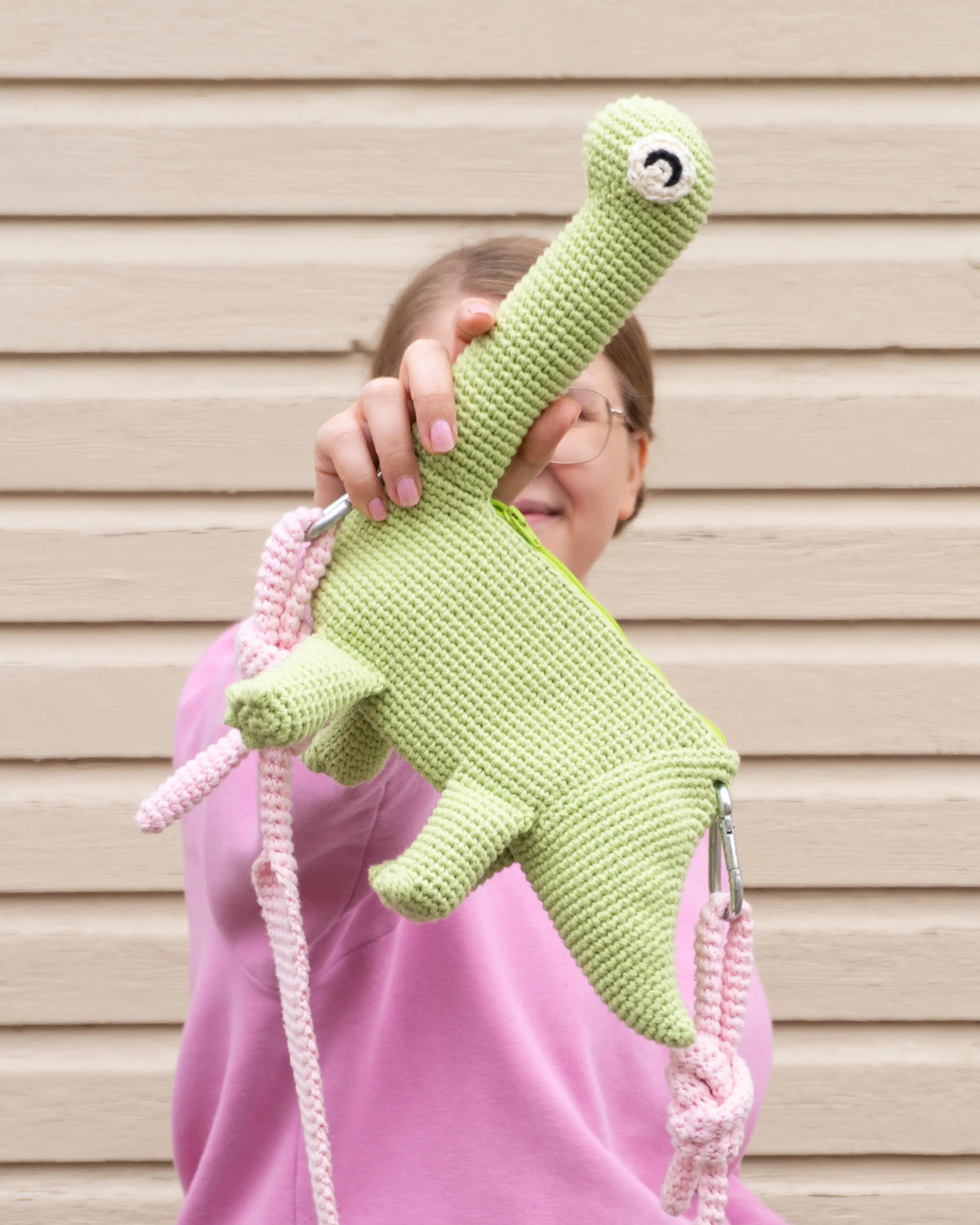 Person holding a green crocheted brachiosaurus dinosaur bag and a pink hoodie.