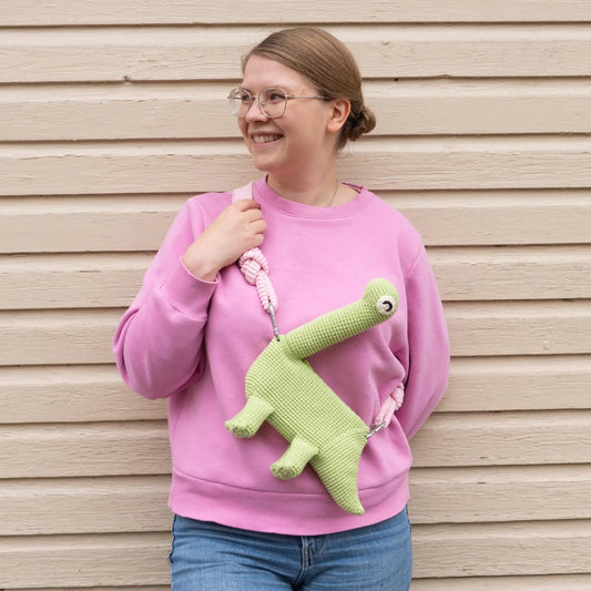 Person wearing a green crocheted brachiosaurus dinosaur bag and a pink hoodie.