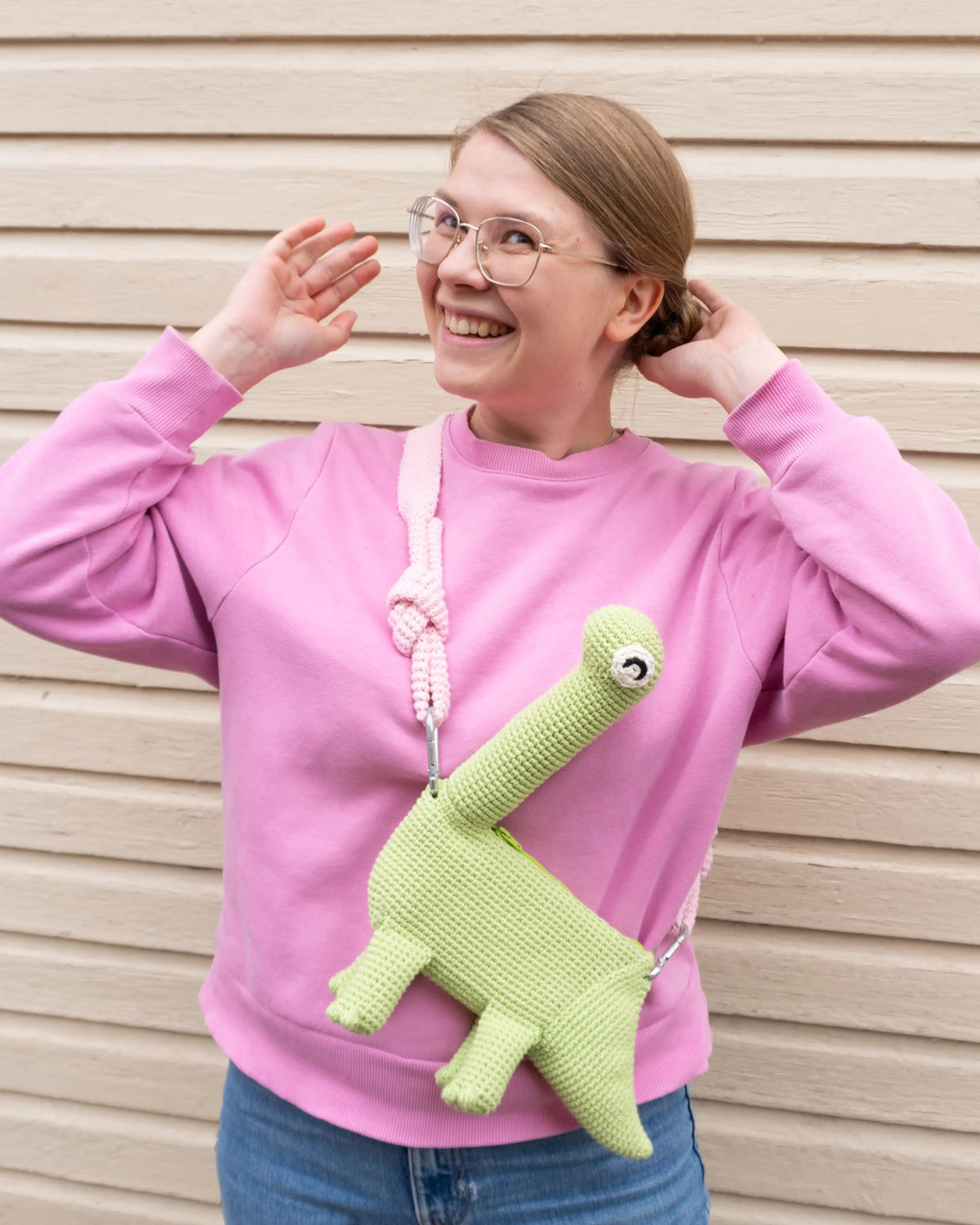 Person wearing a green crocheted brachiosaurus dinosaur bag and a pink hoodie.